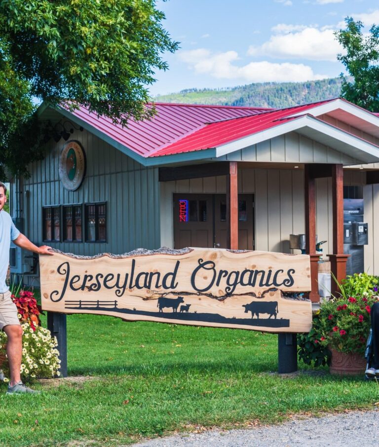Two people stand beside a large wooden sign reading Jerseyland Organics, an agritourism spot in front of a small building with a red roof, surrounded by green grass, trees, and flowers on a sunny day.
