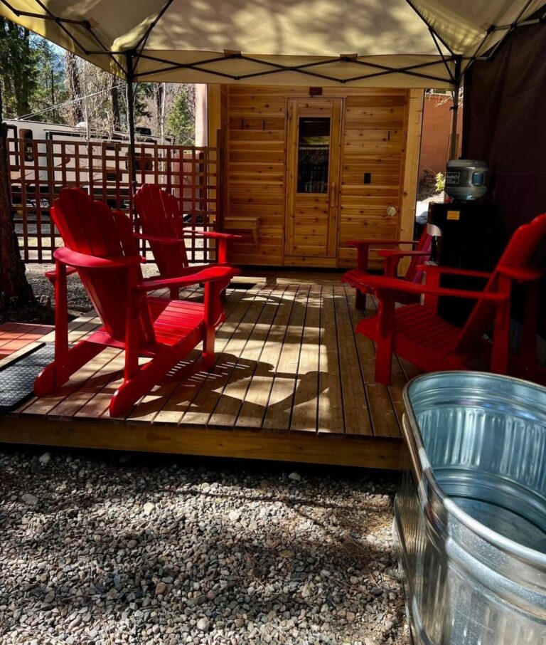 A wooden deck with four bright red Adirondack chairs sits under a canopy in front of a small wooden cabin, offering a peaceful wellness retreat. Nearby, a metal tub and water dispenser invite relaxation amid trees and lattice fencing.