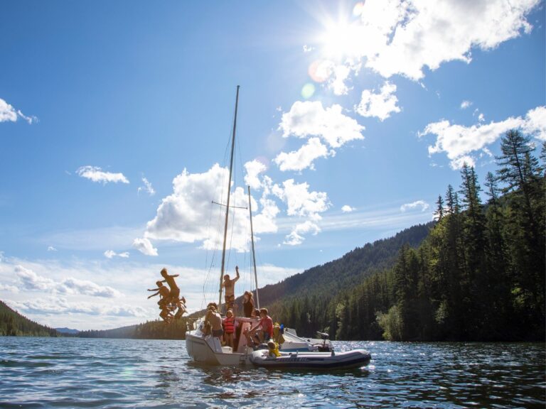 A group of people enjoy a sunny day boating on a sailboat and small inflatable boat on a scenic lake, surrounded by forested hills under a bright blue sky with scattered clouds.