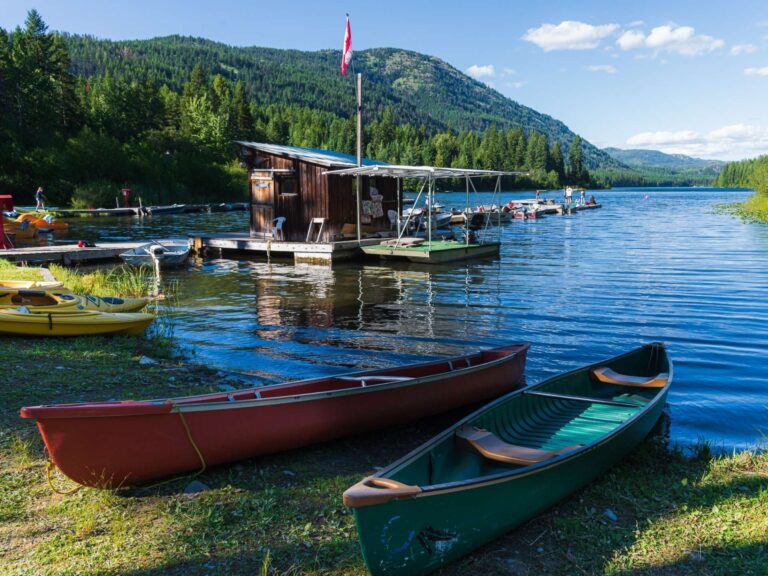 Two canoes rest on grassy shore by a calm lake. A small wooden dock with a Canadian flag and boats are visible on the water, surrounded by forested mountains under a blue sky with scattered clouds.