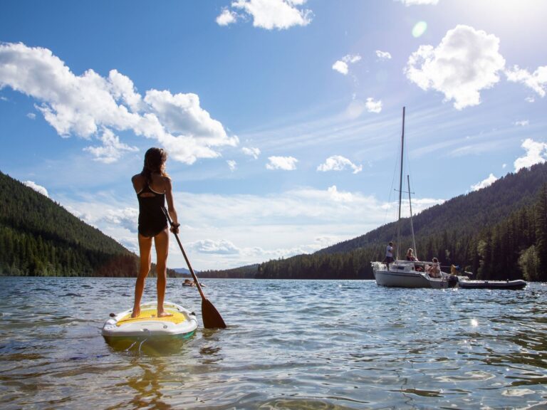 A person stands paddling on a paddleboard in a scenic lake surrounded by forested hills; nearby, a sailboat with people on board floats under a bright, partly cloudy sky.