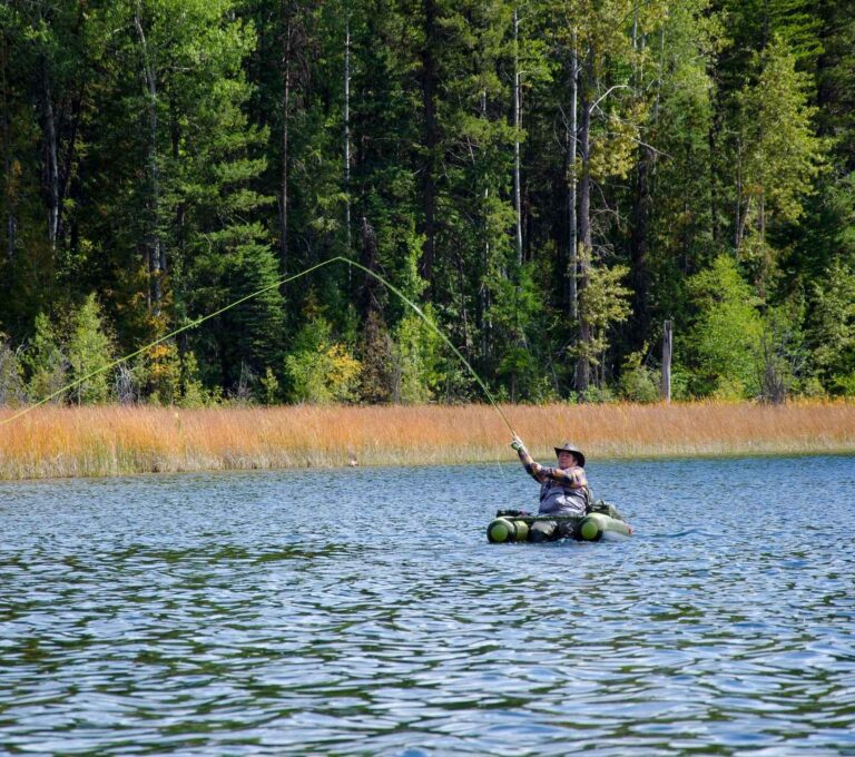 A person wearing a hat and fishing gear enjoys fly-fishing from a small inflatable raft on a calm lake, surrounded by tall trees and grassy shoreline in the background.
