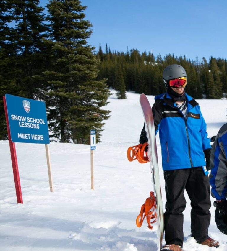 A snowboarder in blue jacket and helmet stands in snow near a “Snow School Lessons Meet Here” sign, with trees and a snowy hill in the background.