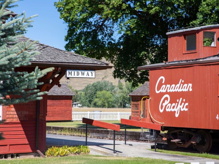 A vintage red Canadian Pacific train car sits on tracks near a wooden building with a Midway sign, surrounded by trees, a fence, and distant hills—capturing the rich history of the area on a sunny day.