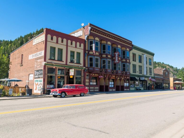 A red vintage car is parked on a quiet street lined with historic brick buildings under a clear blue sky in a small town, creating a perfect scene for photography enthusiasts.