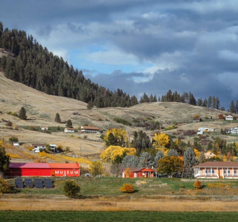 A rural landscape features rolling hills, scattered houses, autumn trees, a red barn labeled MUSEUM highlighting local history, and a large building, all under a dramatic cloudy sky.