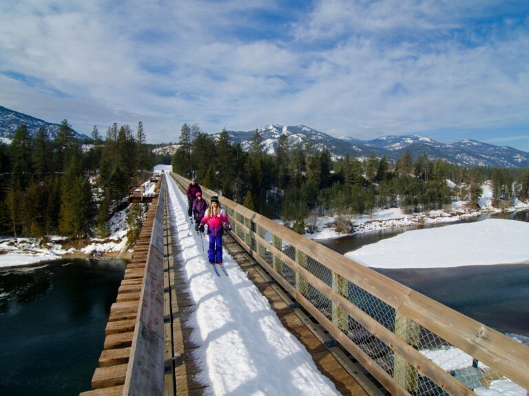 A group of people, including children, walk on a snow-covered wooden bridge over a river with snow and ice, surrounded by pine trees and mountains; some carry Nordic ski gear under a partly cloudy sky.