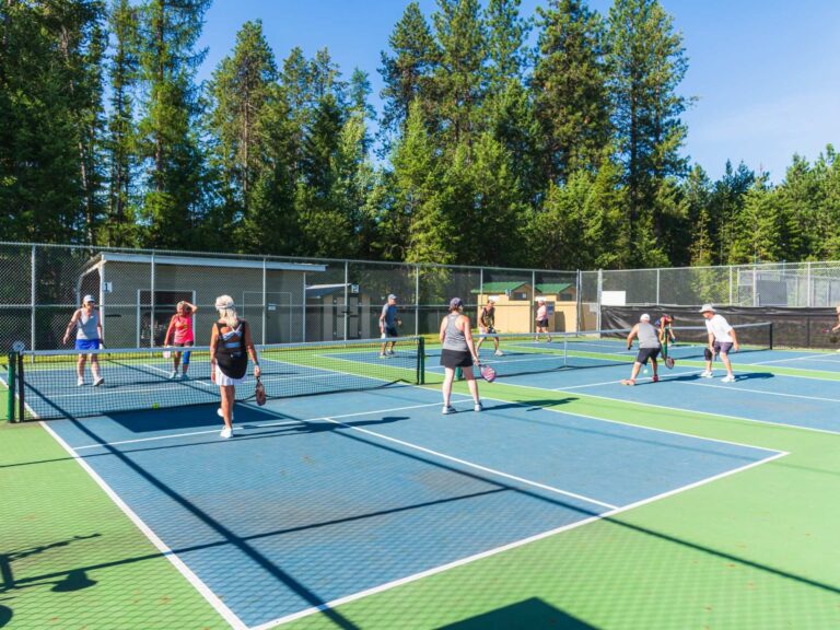 A group of people enjoy a lively game of pickleball on outdoor courts surrounded by tall trees, under a clear blue sky. Some players are near the nets while others wait to return the pickleball.
