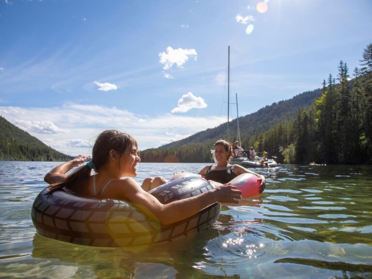 Two people enjoy a river float on inflatable tubes in a clear lake surrounded by forested mountains under a sunny blue sky, with a few others and sailboats visible in the background.
