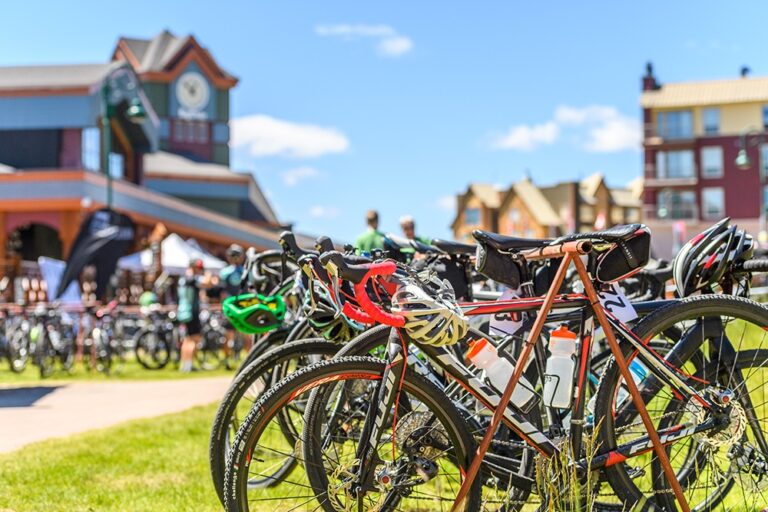 A row of bicycles with helmets hanging on handlebars is parked on a grassy area at Big White, with people and colorful buildings in the background on a sunny day.