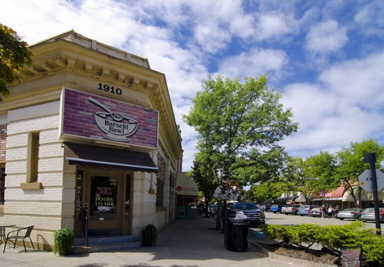 A corner restaurant named Borscht Bowl with a sign and a canopy sits on a sunny, tree-lined street with parked cars and shops. The building, showcasing its history with 1910 engraved near the roof, stands under a partly cloudy sky.