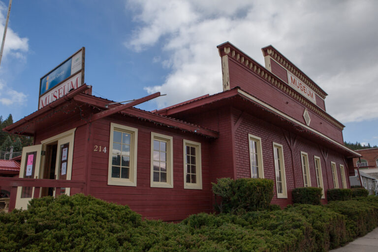 A red wooden building with cream trim houses a history museum, as indicated by signs on the roof and side. The rustic structure is surrounded by green bushes under a partly cloudy sky.