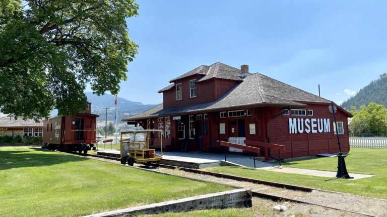 A historic red wooden building marked Museum sits beside old train tracks, showcasing history with a vintage railcar and maintenance trolley nearby, surrounded by green grass and trees on a sunny day.