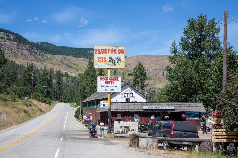 A rural roadside scene with a hotel, pub, and restaurant next to a curving road. Signs read “Prospector Pub” and “Rock Creek Hotel.” Agritourism draws visitors; vehicles and motorcycles are parked outside with mountains and trees in the background.