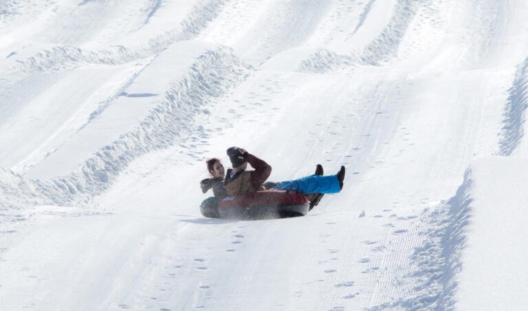 Two people are riding together on an inner tube down a snowy, groomed hill, surrounded by snow-covered tracks under bright sunlight.