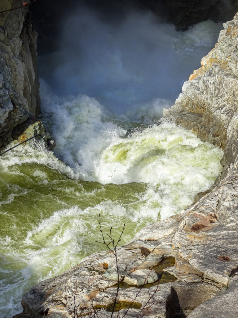 Churning white water rushes powerfully between rocky cliffs, creating mist at the base of a narrow gorge—a dramatic contrast to the peaceful landscapes often found on agritourism adventures nearby. Exposed rocks emphasize the river’s turbulence and force.