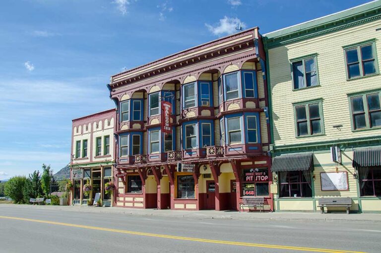 A historic, ornate building with large bay windows and decorative trim stands on a quiet street under a blue sky, showcasing its rich history. The ground floor features businesses, including PIT STOP and a hanging hotel sign.