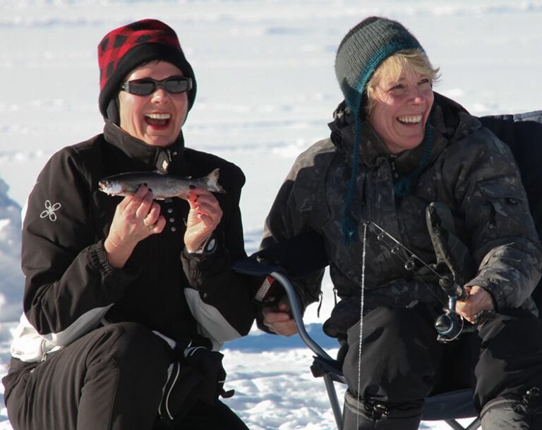 Two smiling women dressed in winter clothing sit on snow while ice fishing. One woman proudly holds a small fish, and the other holds a fishing rod. Both appear happy, enjoying their ice fishing adventure outdoors.
