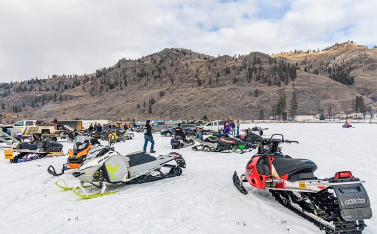 Several snowmobiles are parked on a snowy field with people standing nearby. More snowmobiles and trucks are in the background, while forested hills rise under a partly cloudy sky, creating a classic winter scene for snowmobile enthusiasts.