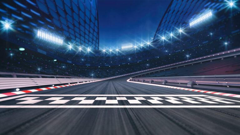 A view of an empty racetrack at night, showing a checkered finish line in the foreground, curved track, stadium lights, and grandstands in the background, creating a dramatic and competitive atmosphere.
