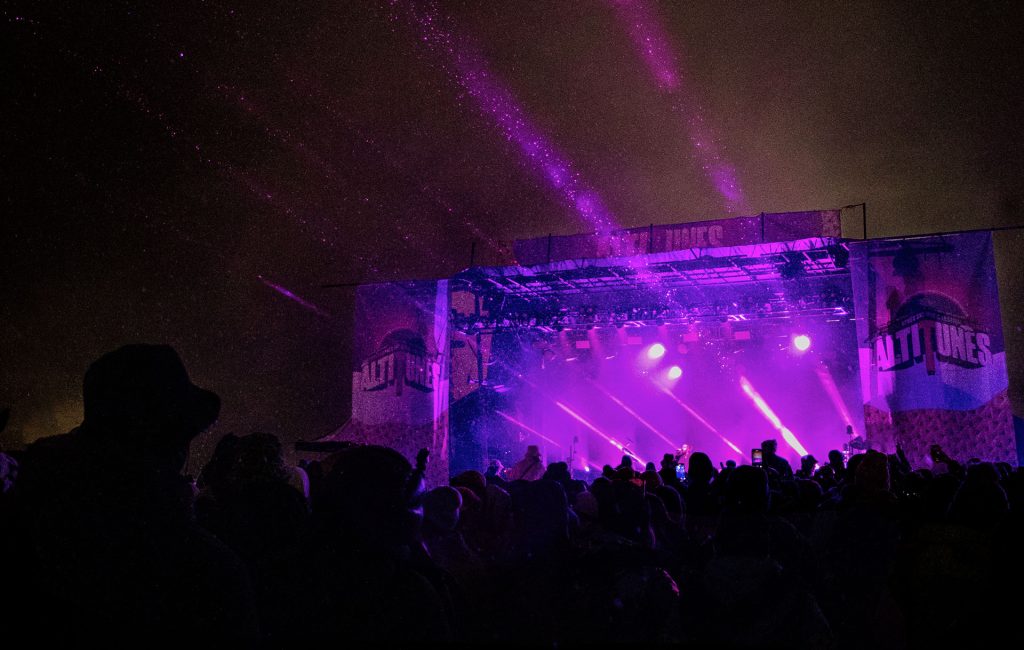 A crowd watches a night concert with purple lights illuminating the stage; the silhouettes of people are visible, and beams of light shine into the dark sky.
