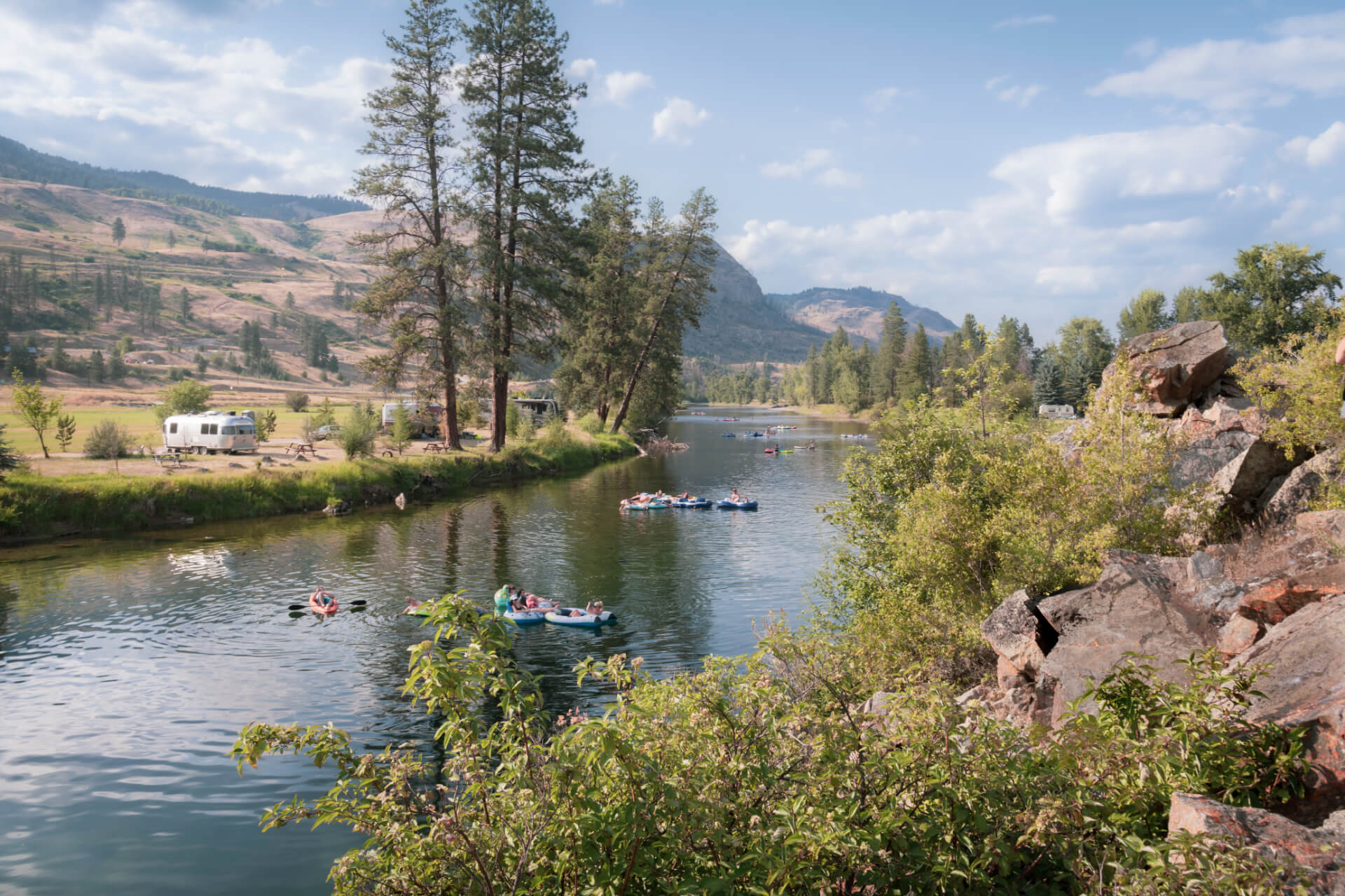 People float on rafts down a calm river in Boundary Country, surrounded by trees and rocky banks, with mountains in the background and a camper trailer parked on the grassy shore under a partly cloudy sky.