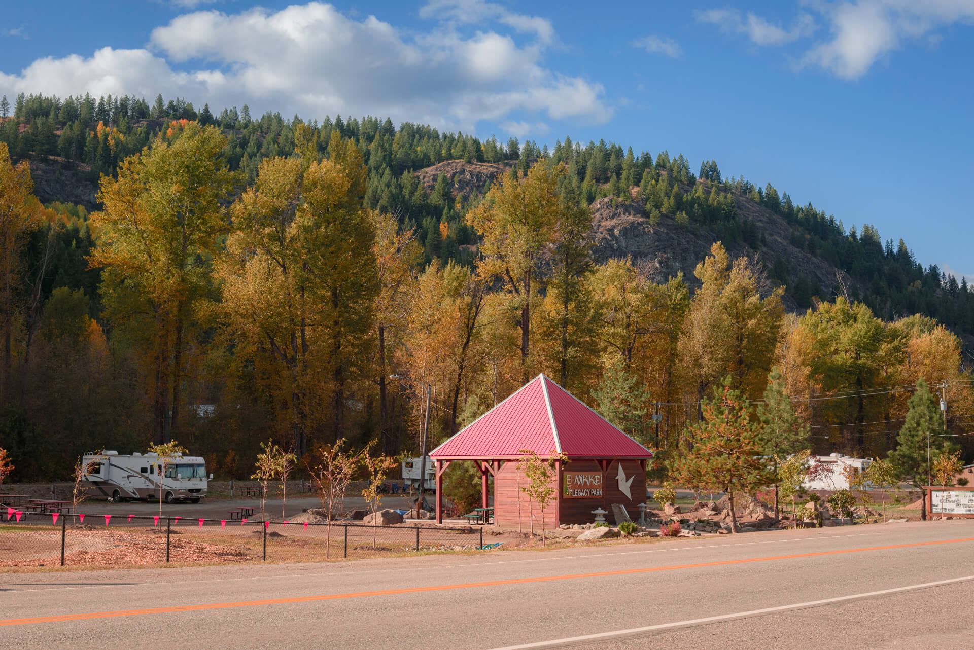 A small pavilion with a red roof stands by a road in Boundary Country, with RVs parked among autumn trees and forested hills in the background under a blue sky with scattered clouds.