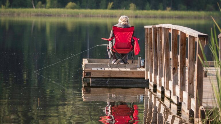 A person with blonde hair sits in a red camping chair on a wooden dock, fishing in a calm lake surrounded by green trees and grass, with their fishing line cast into the water.