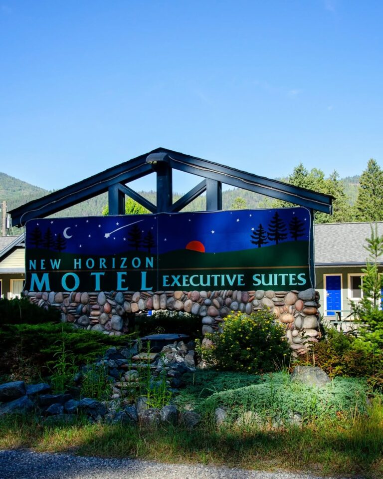 A sign for New Horizon Motel Executive Suites stands in front of a building, with trees, greenery, and mountains in the background under a clear blue sky. The sign features a night sky and sunset illustration, inviting accessible travel for all guests.