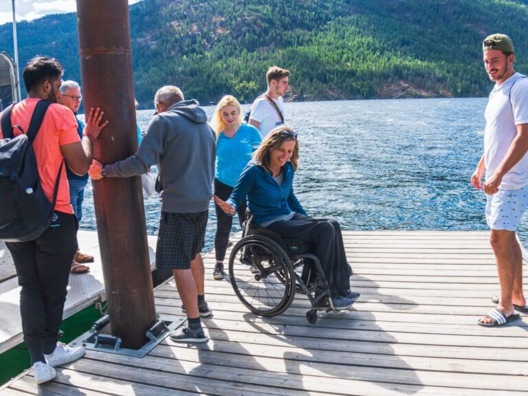 A group of people, including a woman in a wheelchair, gather and interact on a wooden dock by a lake with forested hills in the background, sharing smiles and 7 affirmations. The scene is bright and lively.