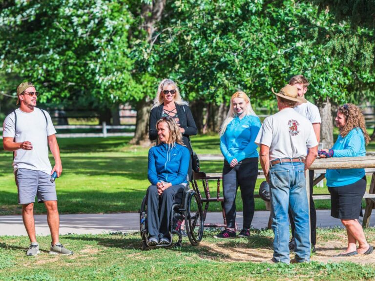 A group of six people, including a woman in a wheelchair, gathers outside on a sunny day, chatting and smiling near trees and a wooden fence in a park, sharing 7 affirmations to uplift each other.