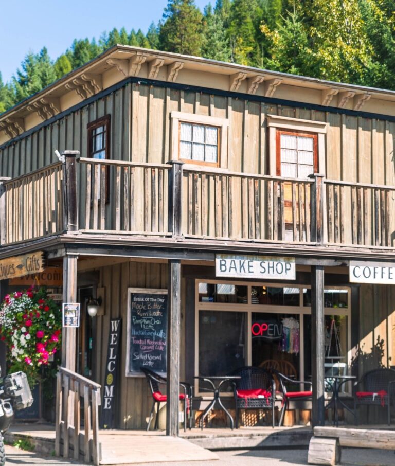 A rustic wooden bake shop and coffee house with a porch, outdoor seating, colorful flower baskets, and a chalkboard menu, set against a backdrop of green trees and sunlight.
