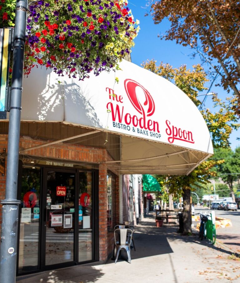 The Wooden Spoon Bistro & Bake Shop storefront with a white awning, brick exterior, and hanging flower basket on a sunny day. Trees with autumn leaves and outdoor seating are visible nearby.