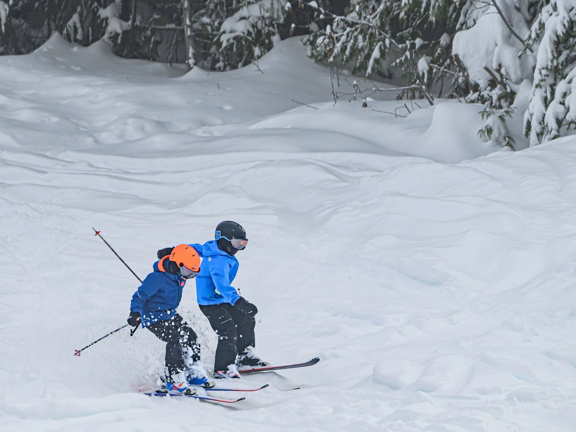 Two children in blue jackets and helmets ski downhill together on a snowy slope surrounded by snow-covered trees.