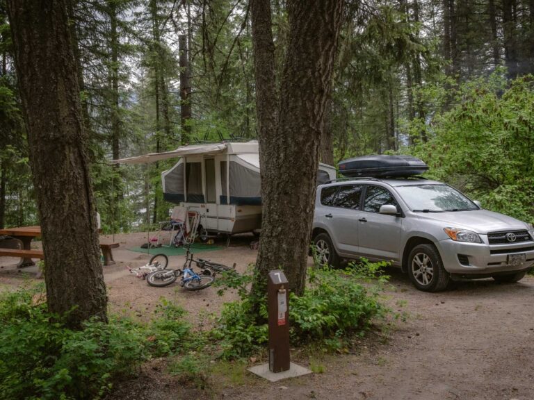A silver SUV is parked next to a pop-up camper in a wooded campsite. A picnic table, two bicycles, and camping gear are nearby, surrounded by tall trees and dense green foliage.