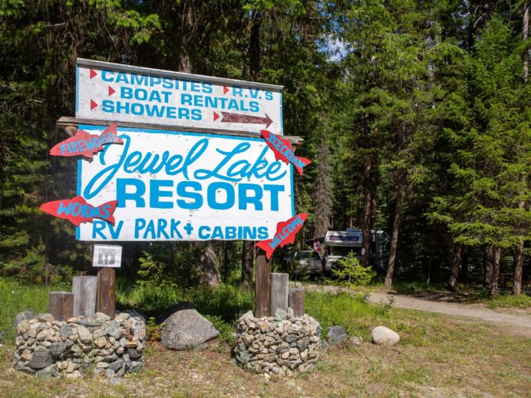 A large sign for Jewel Lake Resort, advertising campsites, RVs, boat rentals, showers, RV park, and cabins. Red arrows point to firewood, restrooms, and a store, surrounded by trees and a gravel path.