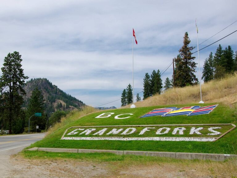 A hillside sign made of white rocks spells out “BC GRAND FORKS” next to a painted British Columbia flag. Two flagpoles stand nearby, with trees, mountains, and a partly cloudy sky in the background.