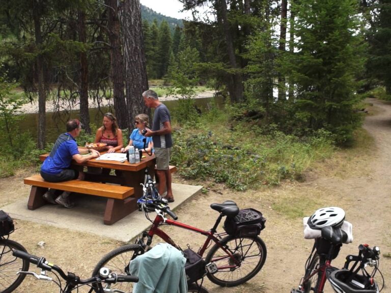 Four people sit at a picnic table in a forested area near a river, talking and eating. Bicycles with helmets and gear are parked nearby on a dirt path leading into the trees.