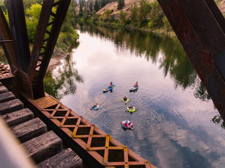 A group of people float on inner tubes and paddleboards in a calm river, framed by the beams of an old railroad bridge, with trees and a hillside in the background.