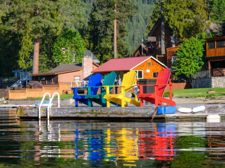 Four colorful Adirondack chairs (blue, green, yellow, and red) sit on a dock by a lake, reflecting in the water. Behind them are houses, greenery, and tall trees on a sunny day.