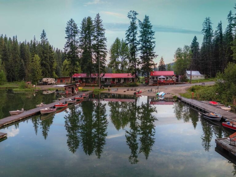 A calm lake reflects nearby pine trees and a row of red-roofed cabins. Small boats are docked along wooden piers, and a sandy shore leads to the cabins surrounded by a forest. The scene is tranquil and serene.