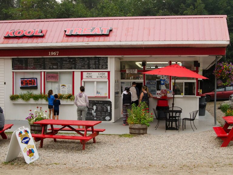 A small roadside ice cream stand with a red roof and sign reading Kool Treat. People are ordering at the window. Red picnic tables and a red umbrella are outside, and a menu board is visible above the counter.
