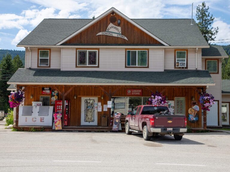 A red pickup truck is parked outside a rustic two-story general store with wood siding and flower baskets. A large ice freezer and vending machines are visible near the entrance.