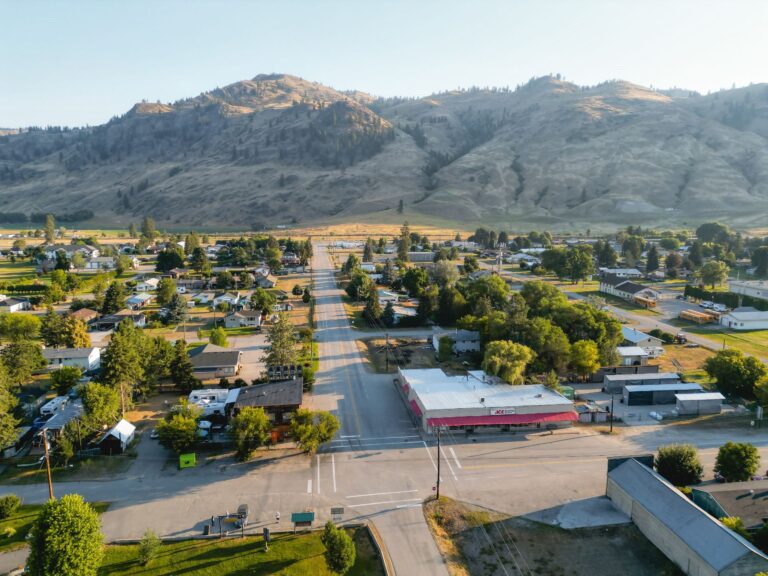 Aerial view of a small town with a main street, residential houses, green trees, and hills in the background under clear skies. The streets appear quiet and the town is surrounded by natural landscape.