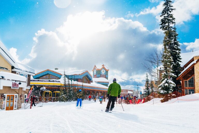 A skier in a green jacket glides down a snowy path toward a ski resort village with colorful buildings and a clock tower, under a bright, partly cloudy sky. Other people are walking and enjoying the winter scenery.