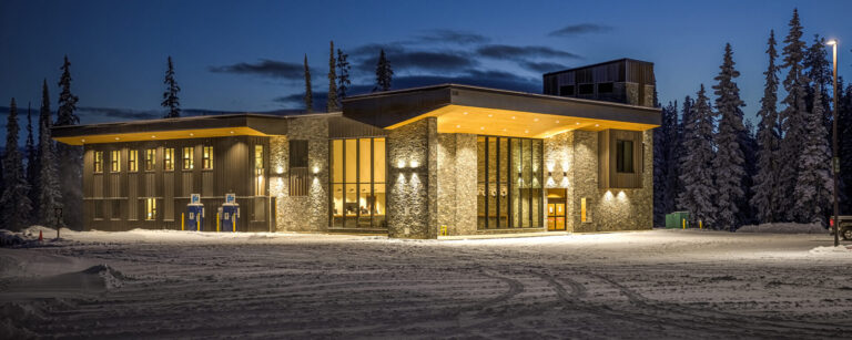 Modern lodge-style building with large windows, lit warmly at dusk, surrounded by snow and tall pine trees under a darkening Boundary Country sky.