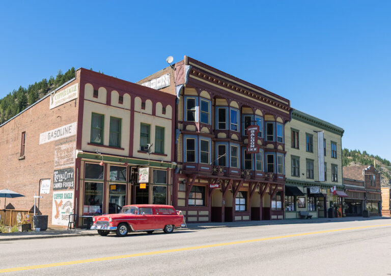 A bright red vintage car is parked on the street in front of colorful, historic western-style buildings, inviting visitors to stay and eat under a clear blue sky in this charming small town setting.