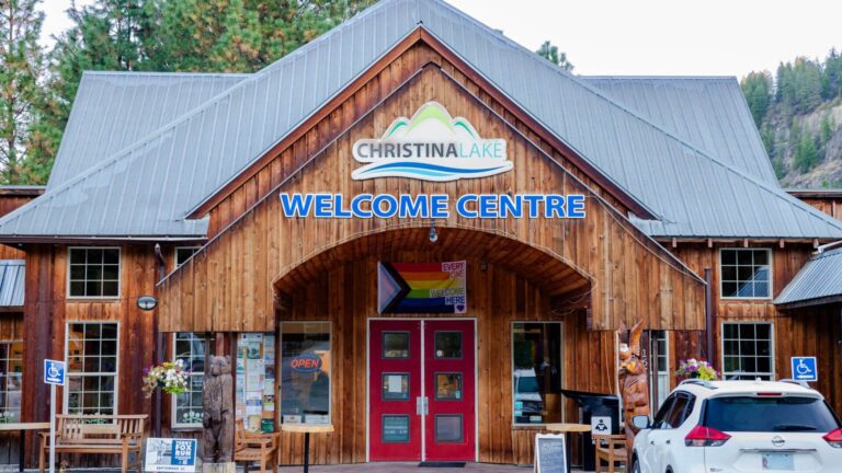 A wooden building with a sign reading “Christina Lake Welcome Centre” above the entrance serves as one of the inviting Visitor Centres. A white car is parked nearby, flowers decorate the entrance, and the building is set among trees and hills.