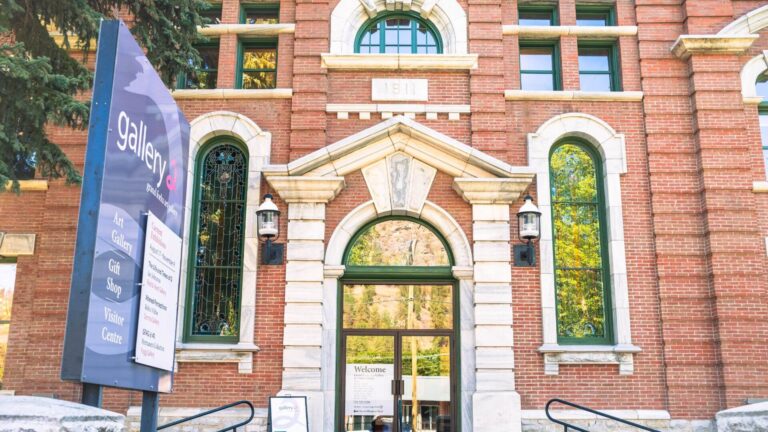 Historic red-brick building with arched windows, ornate stone trim, and a central entrance. A sign in front highlights Visitor Centres, providing directions to the art gallery, gift shop, and visitor centre. Trees partially frame the building.