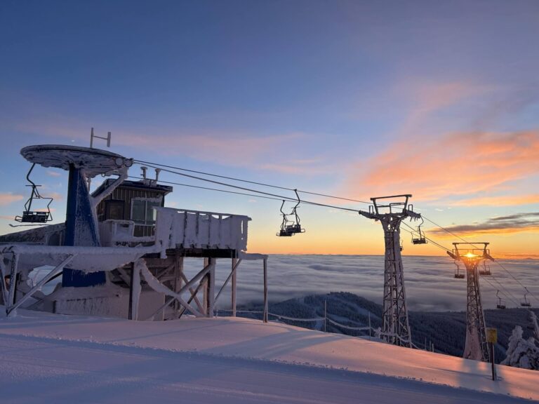 A snow-covered ski lift station sits atop a Boundary Country mountain at sunrise, with chairlifts hanging over a snowy slope and clouds blanketing the valley below, under a colorful sky.